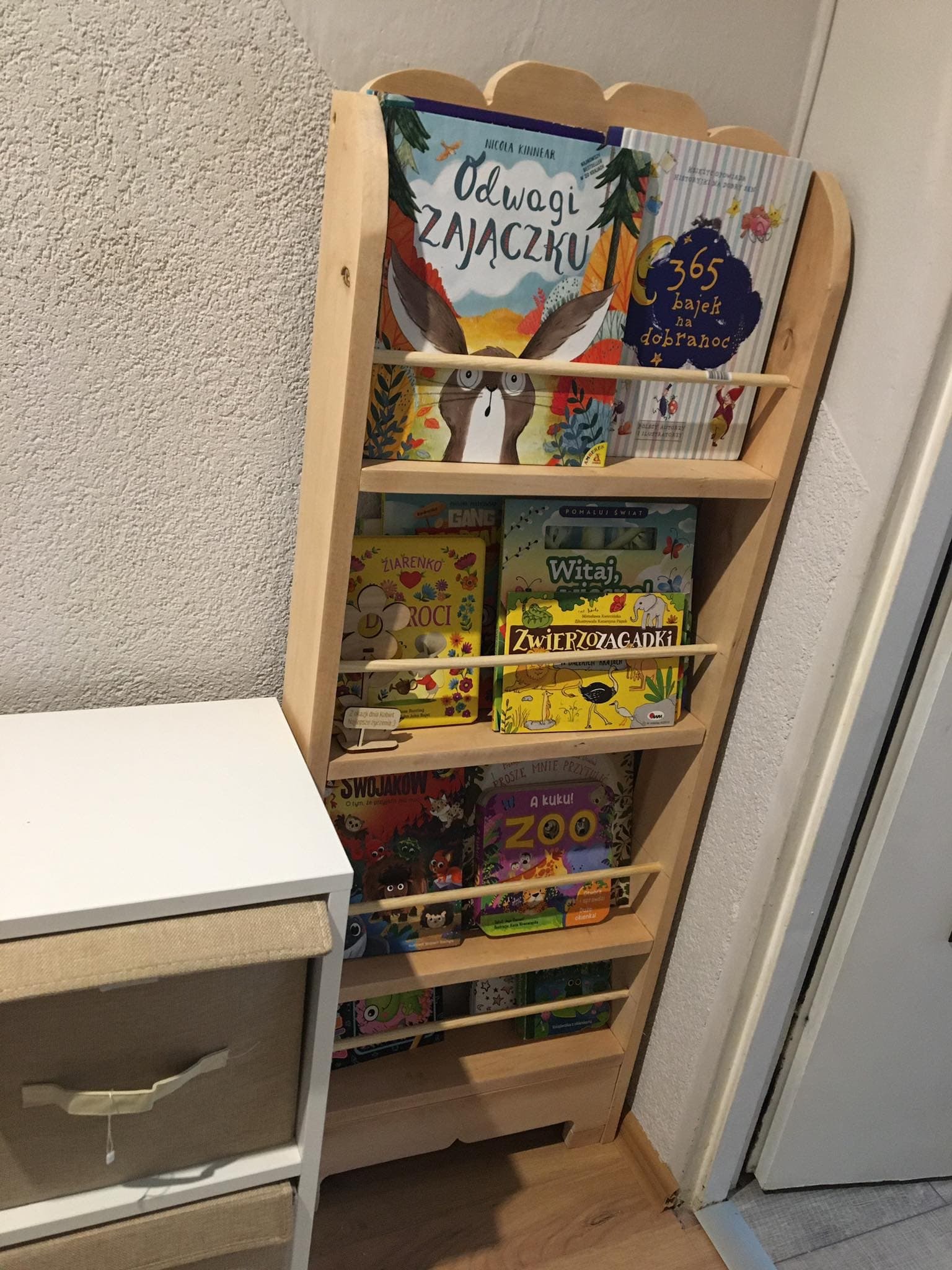 Drewniany regał na książki dla dzieci z zaokrąglonymi krawędziami, stojący w kącie pokoju w Biłgoraju, wypełniony kolorowymi książeczkami z bajkami i opowiadaniami o zwierzętach.