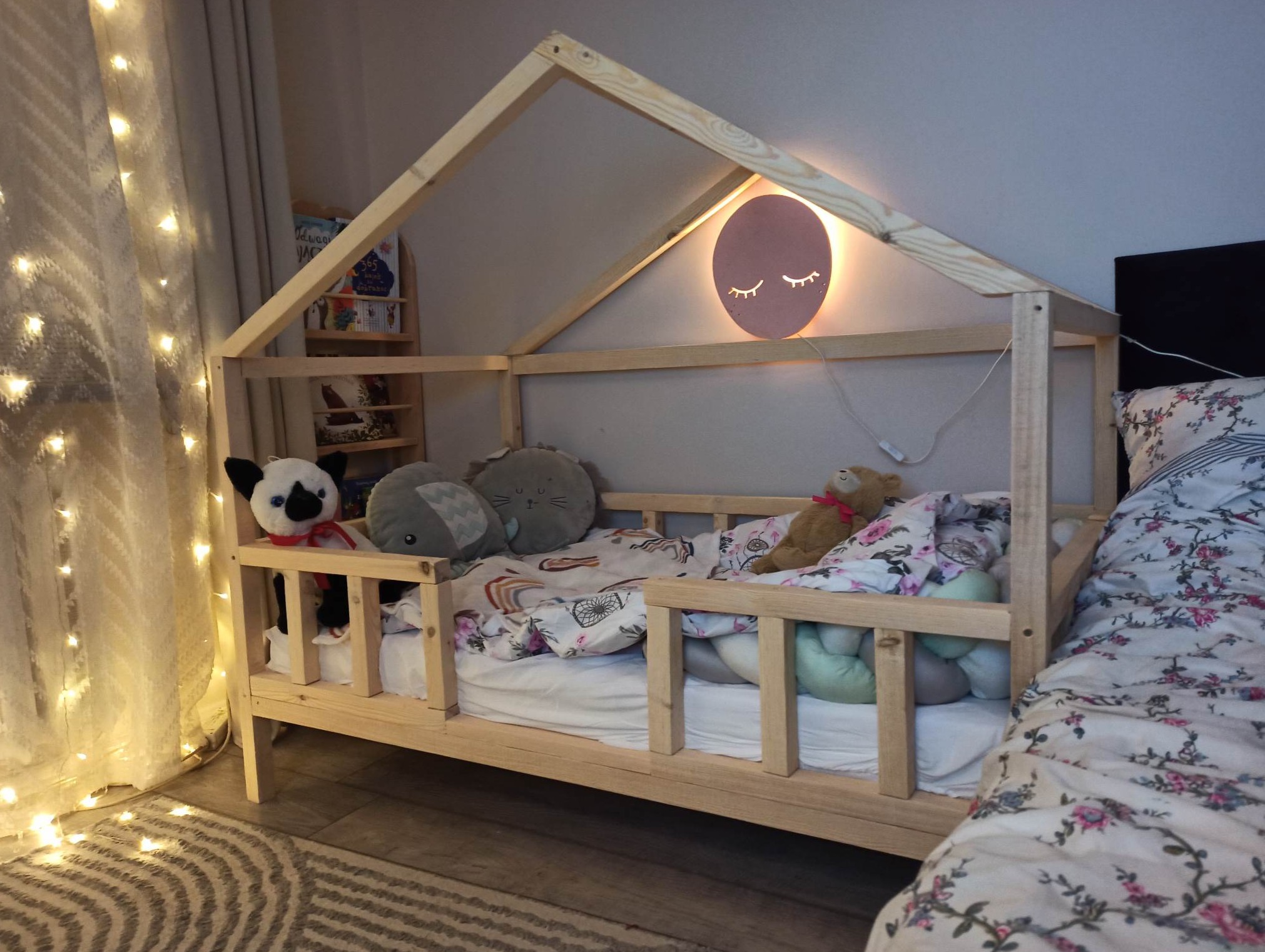 Pokój dziecięcy z łóżkiem w kształcie domku z surowego drewna, ozdobionym lampką nocną w kształcie twarzy i girlandą świetlną, obok stoi łóżko dla rodziców.