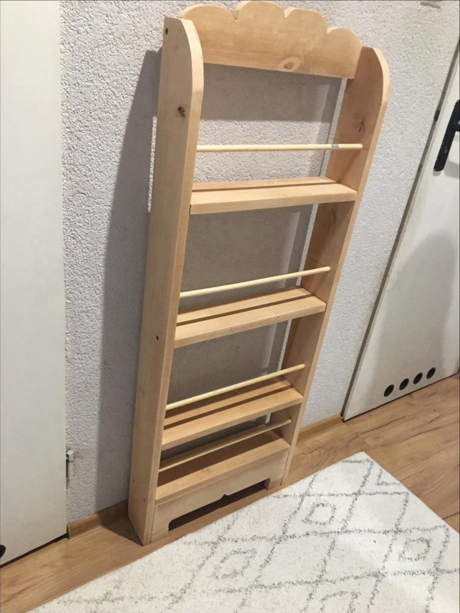 Drewniany regał na książki dla dzieci, z zaokrąglonymi krawędziami i zabezpieczeniami w formie drewnianych prętów, ustawiony na puszystym dywanie w pokoju.