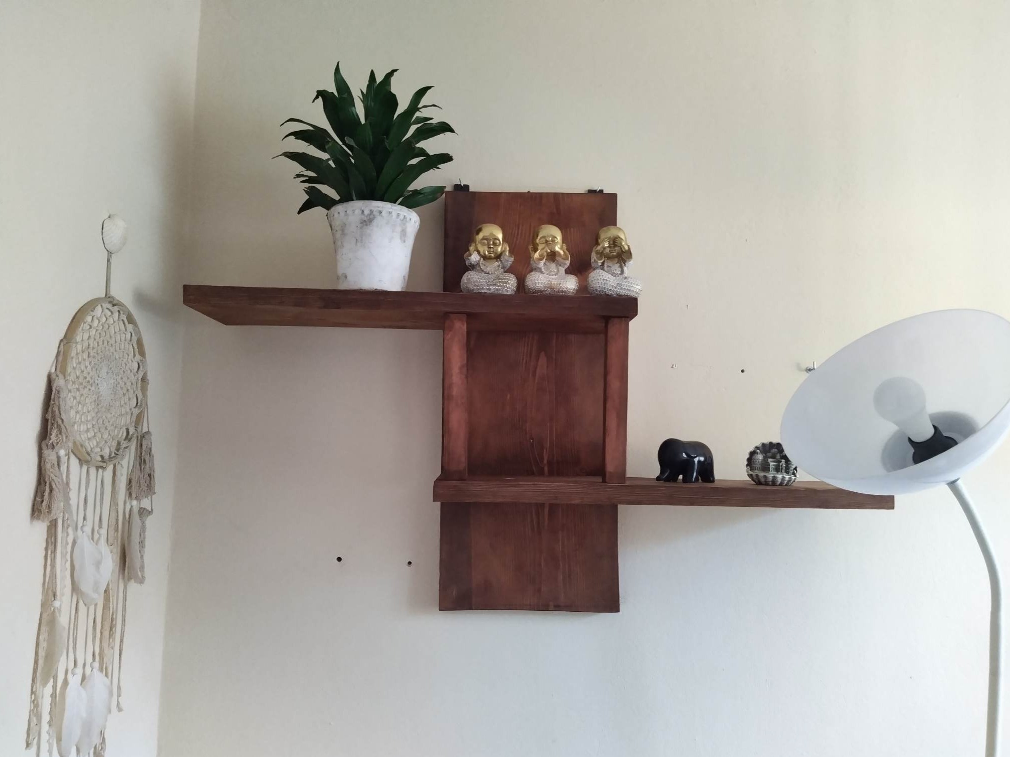 Drewniana półka wisząca w kolorze ciemnego brązu z rośliną doniczkową, trzema figurkami buddy, czarnym słoniem i miniaturą budynku, wisząca na białej ścianie obok łapacza snów i lampy