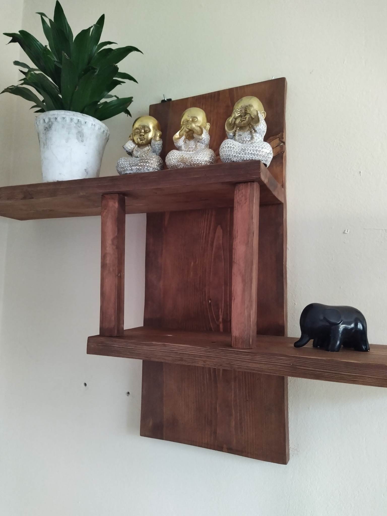 Dwupoziomowa, ciemna drewniana półka ścienna z trzema figurkami Buddy na górnym poziomie i czarną figurką słonia na dolnym poziomie, obok stoi doniczka z zieloną rośliną.