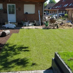 Układanie trawy z rolki w ogrodzie, widoczny walec do ubijania ziemi, mieszalnik do betonu oraz fragment posadzonej trawy w korycie