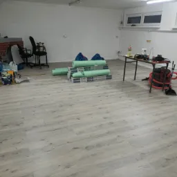 Jasne panele podłogowe imitujące drewno, ułożone w pomieszczeniu z widocznymi materiałami izolacyjnymi, narzędziami i meblami, ukazujące etap prac wykończeniowych.