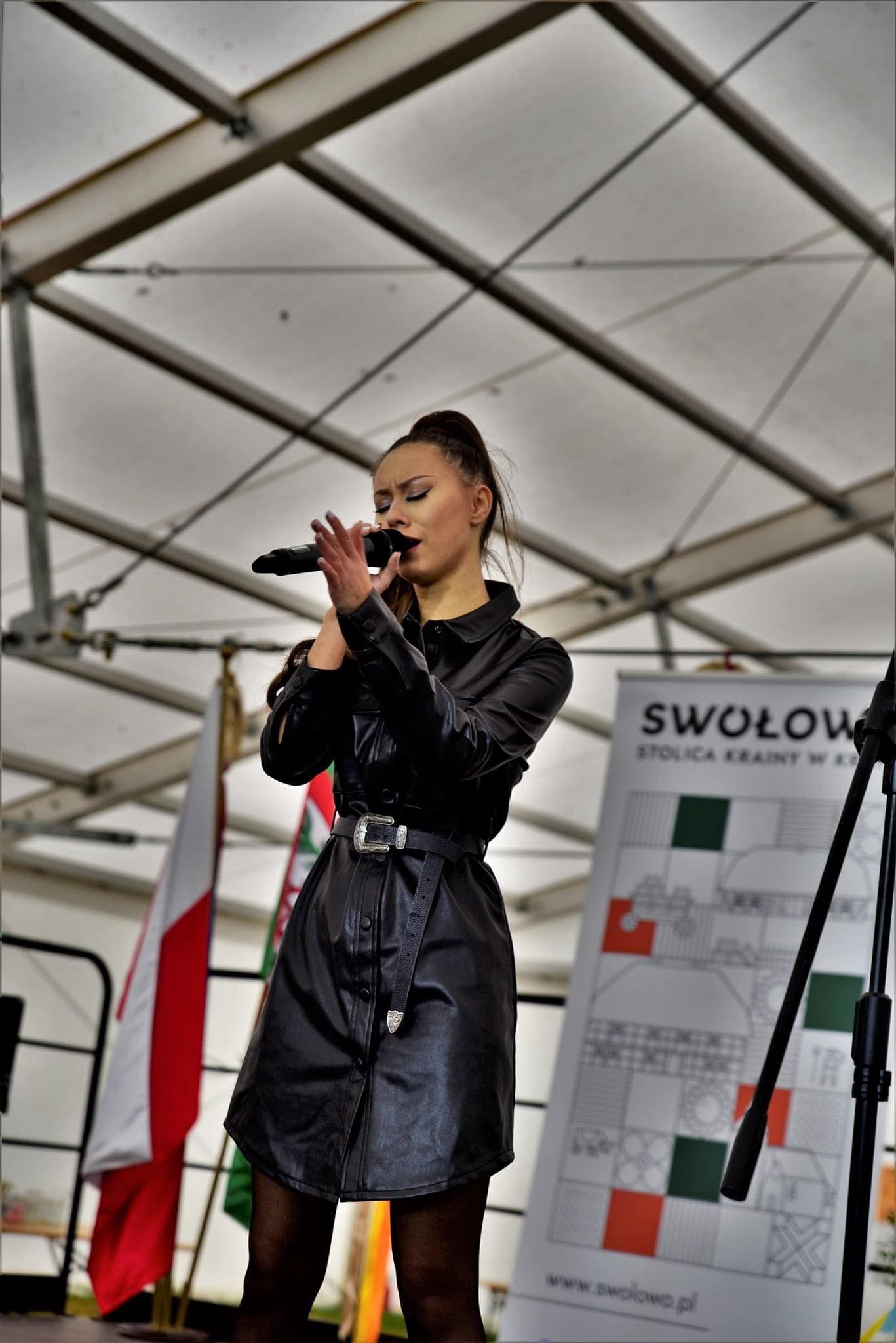 Wokalistka w czarnej, skórzanej sukience śpiewa do mikrofonu na scenie podczas plenerowego występu, z flagami i plakatem w tle.
