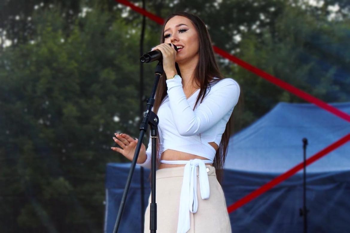 Młoda kobieta z długimi brązowymi włosami śpiewa do mikrofonu na scenie, ubrana w białą bluzkę z długim rękawem i beżową spódnicę z wiązaniem.