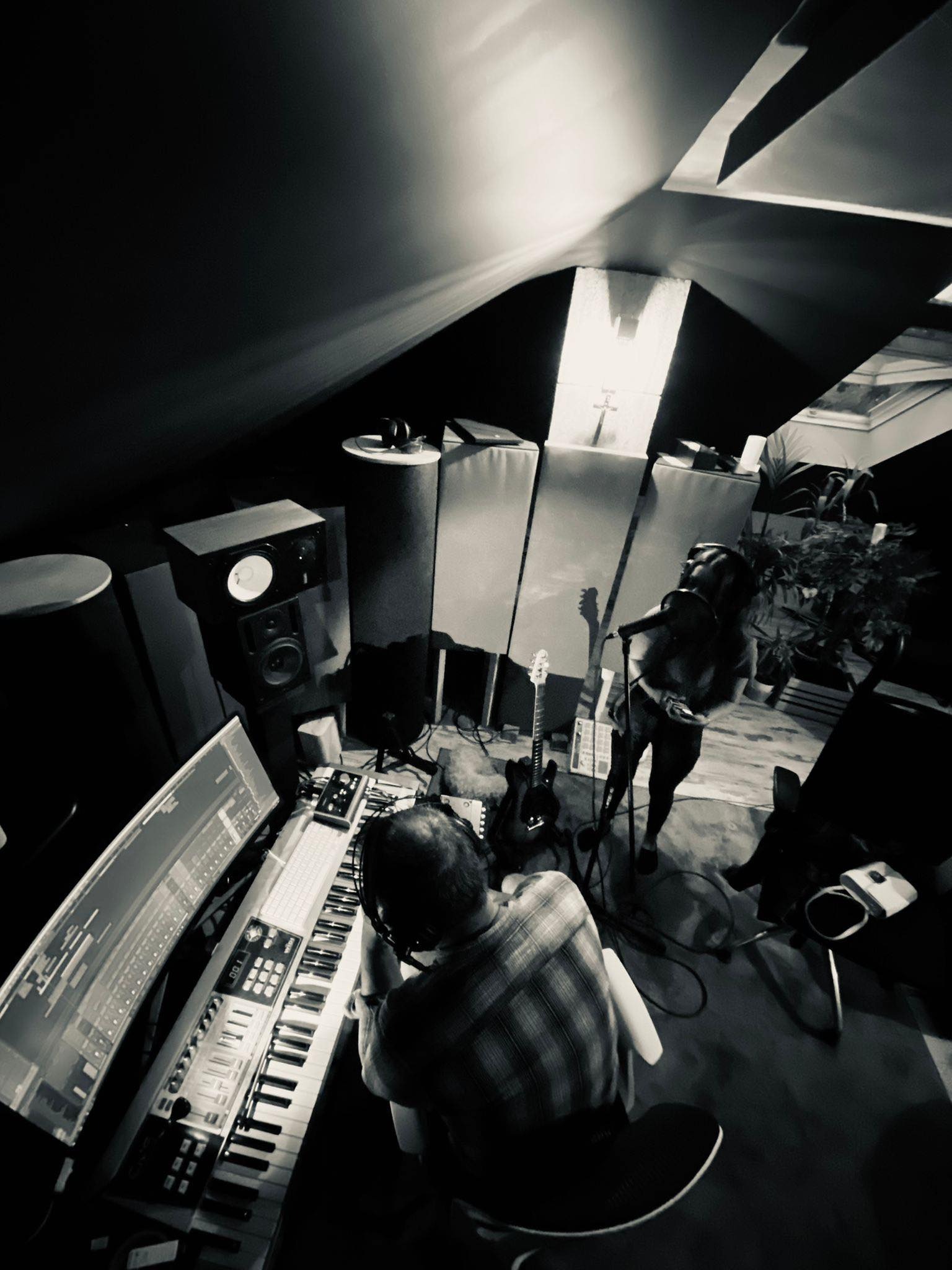 Czarno-białe zdjęcie studia nagraniowego z wokalistką śpiewającą do mikrofonu i mężczyzną obsługującym mikser i klawiaturę, gitara w tle.