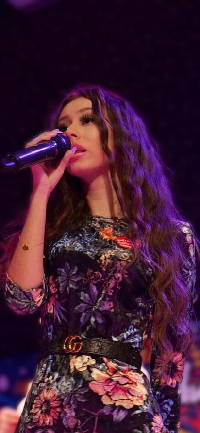 Młoda kobieta z długimi, falowanymi włosami śpiewa do mikrofonu na scenie w fioletowym oświetleniu, ubrana w sukienkę w kwiaty z czarnym paskiem z klamrą w kształcie litery G.