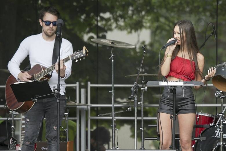 Wokalistka w czerwonym topie i gitarzysta w okularach słonecznych występują na scenie podczas plenerowego koncertu, widoczne perkusja i keyboard.