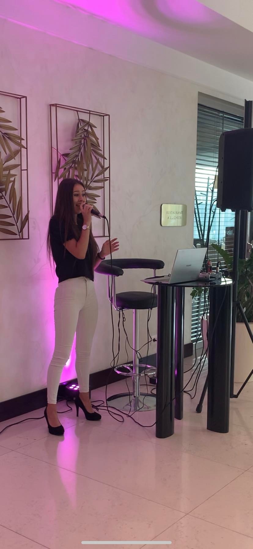 Wokalistka w czarnej bluzce i białych spodniach śpiewa do mikrofonu na tle ściany z dekoracjami roślinnymi i stolikiem z laptopem w lokalu z napisem 'Restaurant A la Carte'.