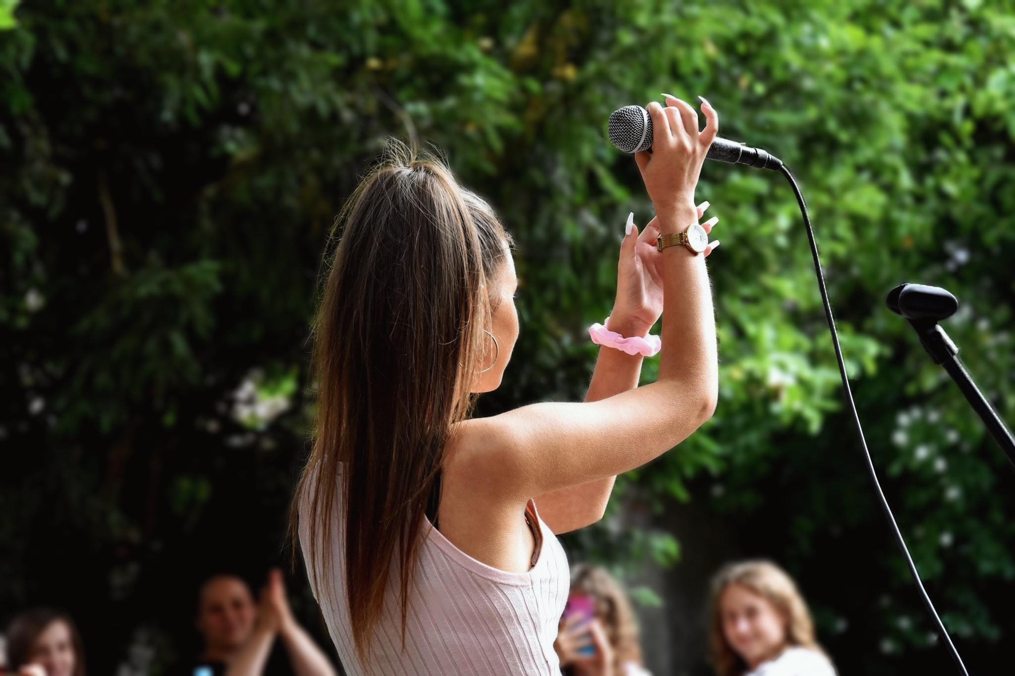 Kobieta z długimi paznokciami trzyma mikrofon na tle zielonych drzew, widoczne osoby w tle.