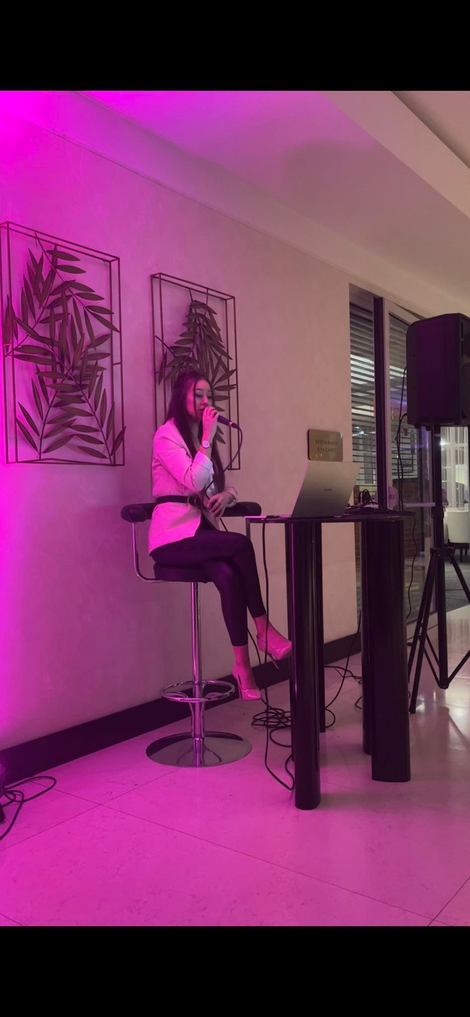 Wokalistka w różowym żakiecie śpiewa do mikrofonu siedząc na wysokim krześle barowym, obok stolik z laptopem i głośnik, dekoracje ścienne z motywem liści, oświetlenie w kolorze fuksji.