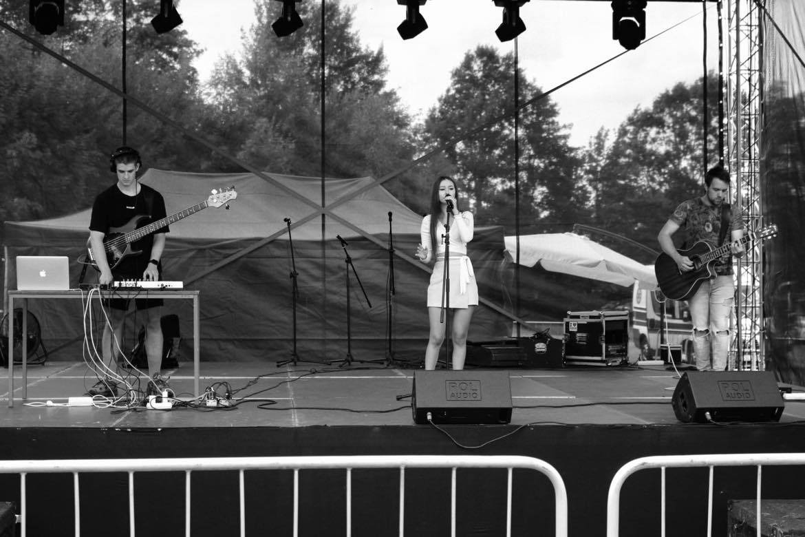 Zespół muzyczny w czerni i bieli na scenie plenerowej: wokalistka w białym stroju, basista z laptopem i gitarzysta z gitarą akustyczną, widoczne reflektory i nagłośnienie.