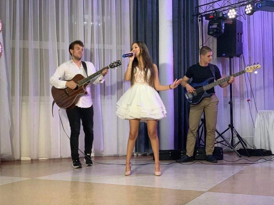 Zespół muzyczny na scenie: wokalistka w białej sukience śpiewa do mikrofonu, gitarzysta gra na gitarze akustycznej, basista gra na gitarze basowej, oświetlenie sceniczne w tle.