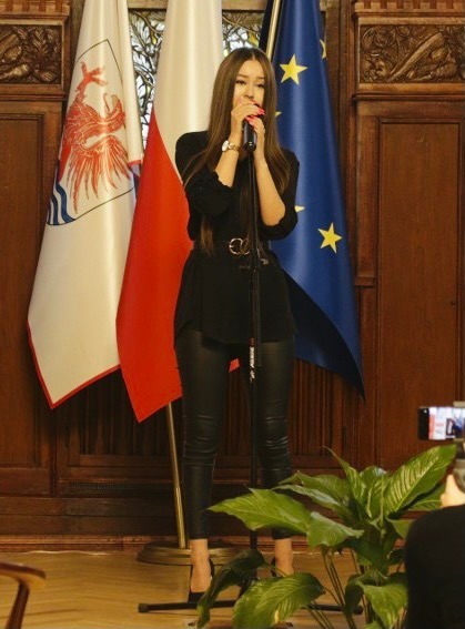 Młoda kobieta w czarnym stroju śpiewa do mikrofonu na tle flagi Polski i Unii Europejskiej, nagrywana telefonem.