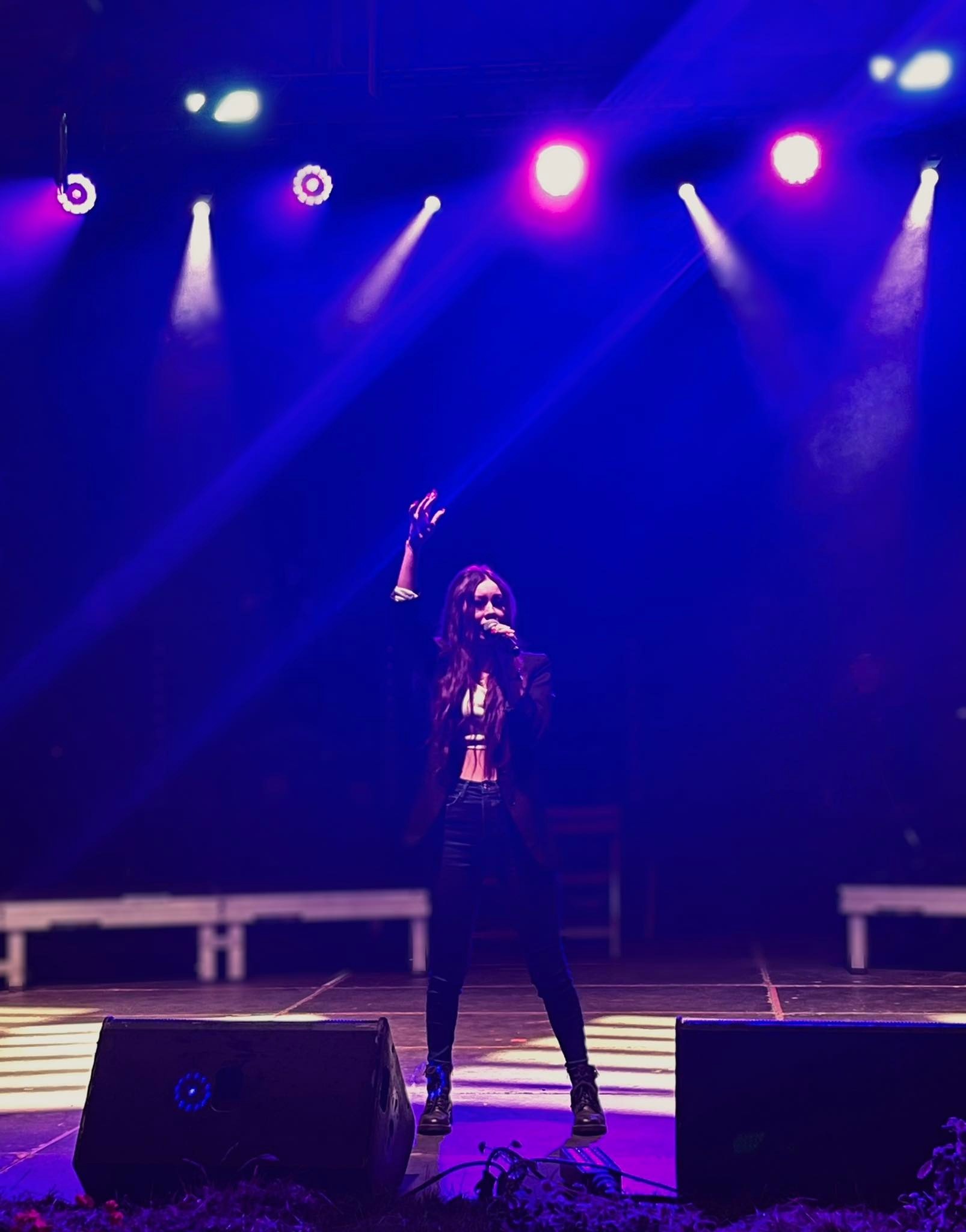Piosenkarka z długimi włosami śpiewa na scenie w blasku niebieskich i różowych świateł, mikrofon w dłoni, podniesiona ręka, widoczne głośniki i elementy scenografii.