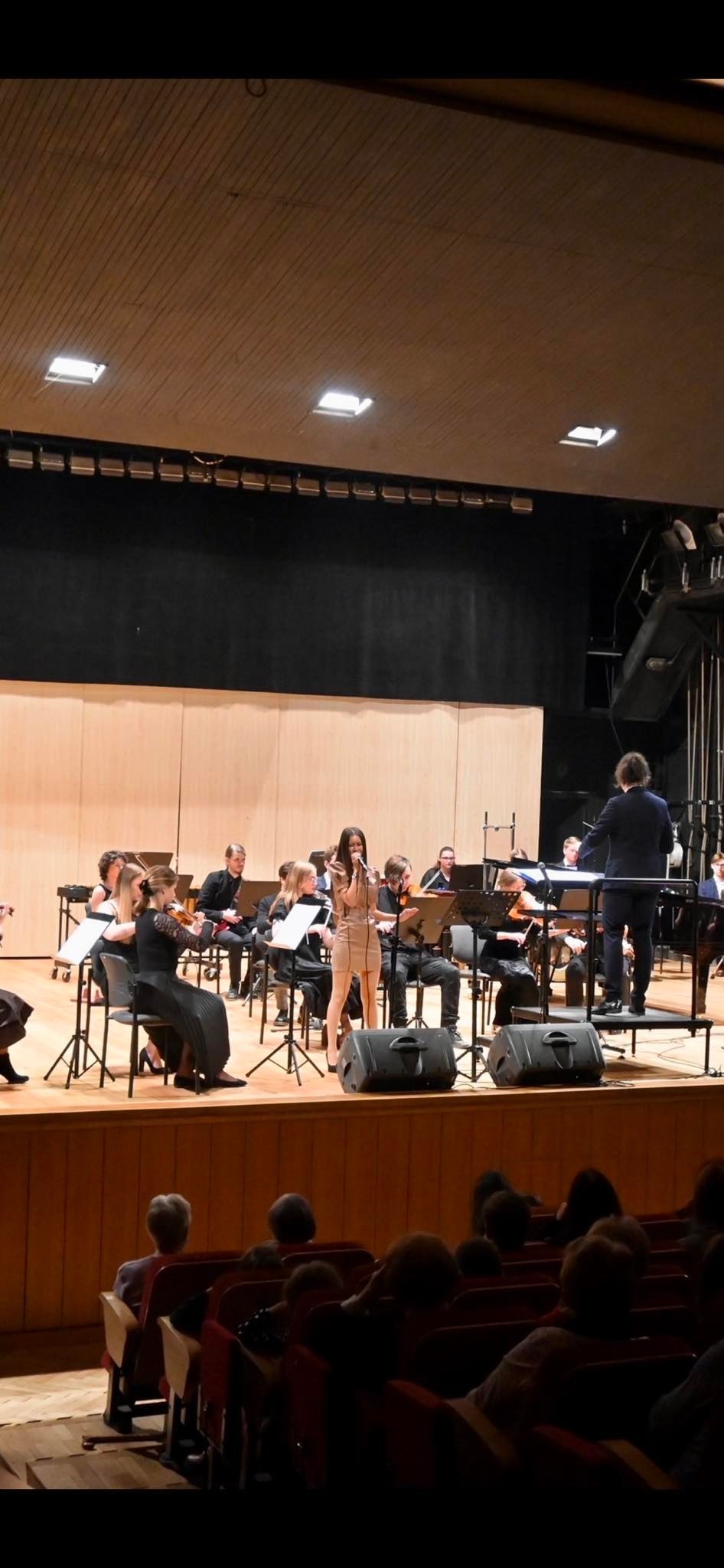 Wokalistka w beżowej sukience śpiewa na scenie z orkiestrą smyczkową i dyrygentem podczas występu dla publiczności.