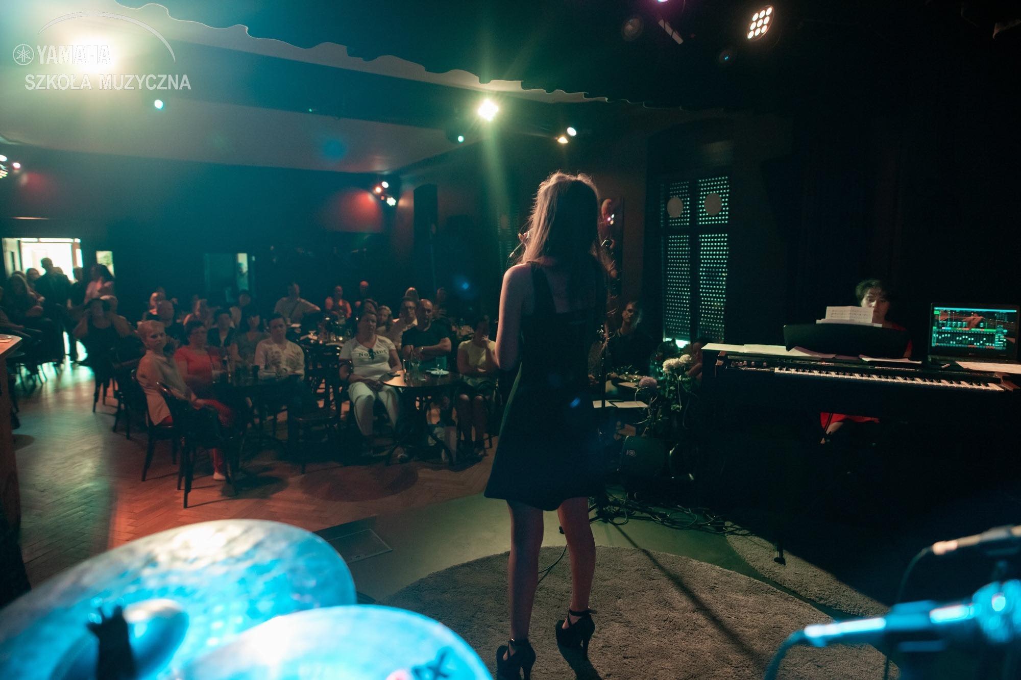Występująca w Słupsku wokalistka na scenie, widziana zza pleców, z widoczną publicznością przy stolikach i pianistą w tle.