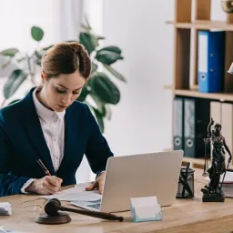 Kobieta w granatowym żakiecie, pisząca na dokumentach przy biurku z laptopem, obok waga sprawiedliwości i młotek sędziowski.