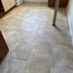 Beżowe, matowe płytki podłogowe ułożone w kuchni, z widocznymi fugami, fragmentem szafki kuchennej i nogą stołu w tle.