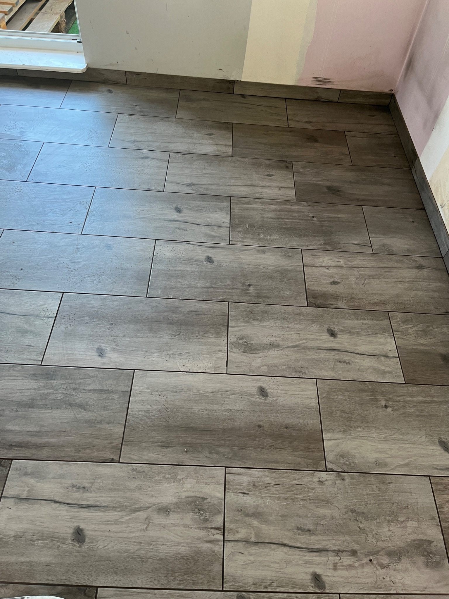 Ułożona podłoga z płytek imitujących drewno w odcieniach szarości, widoczne fugi i fragment ściany z śladami po malowaniu.