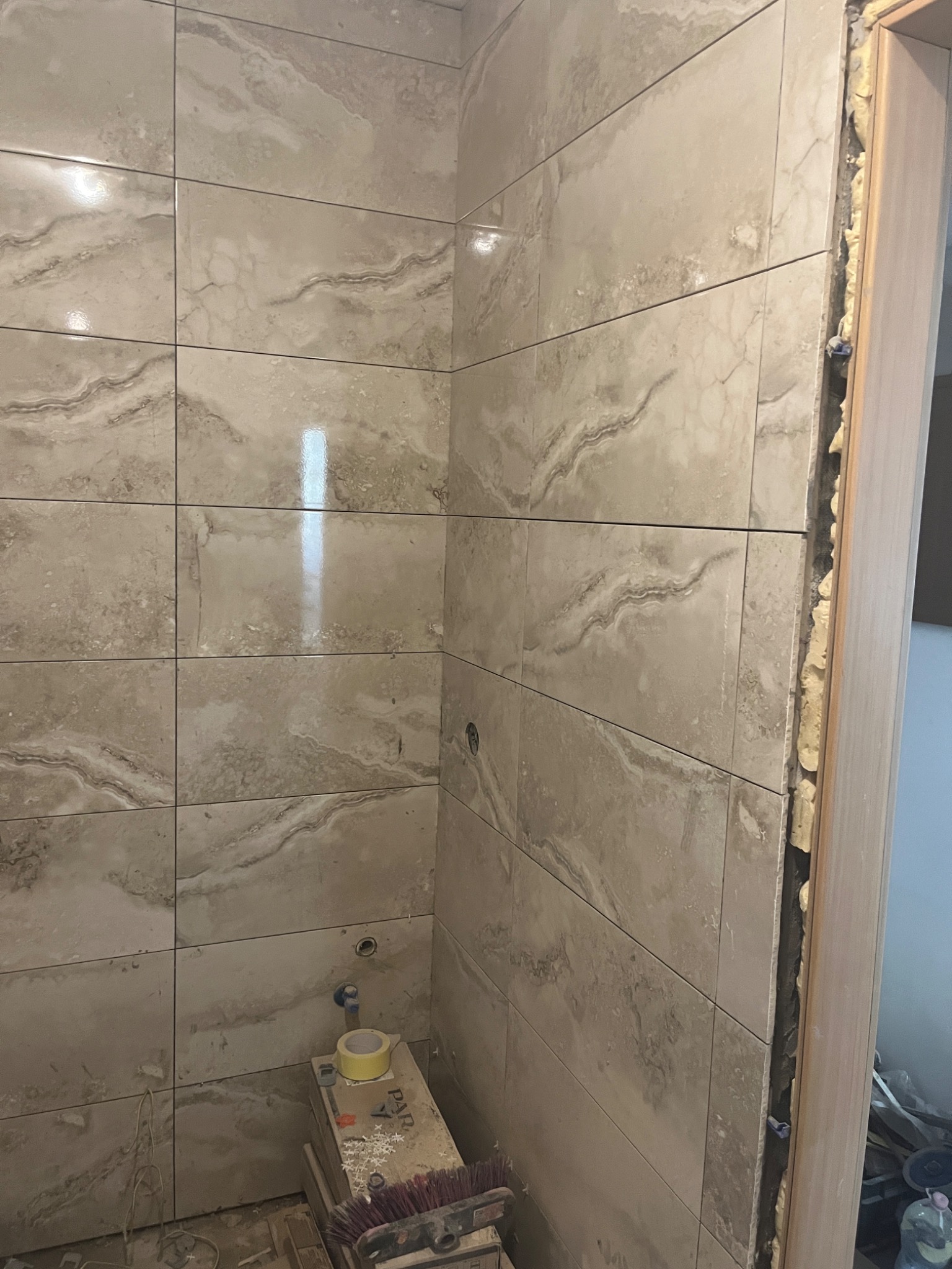 Ściana wyłożona beżowymi płytkami imitującymi marmur, widoczne narzędzia glazurnicze i resztki po fugowaniu, pianka montażowa wokół ościeżnicy.