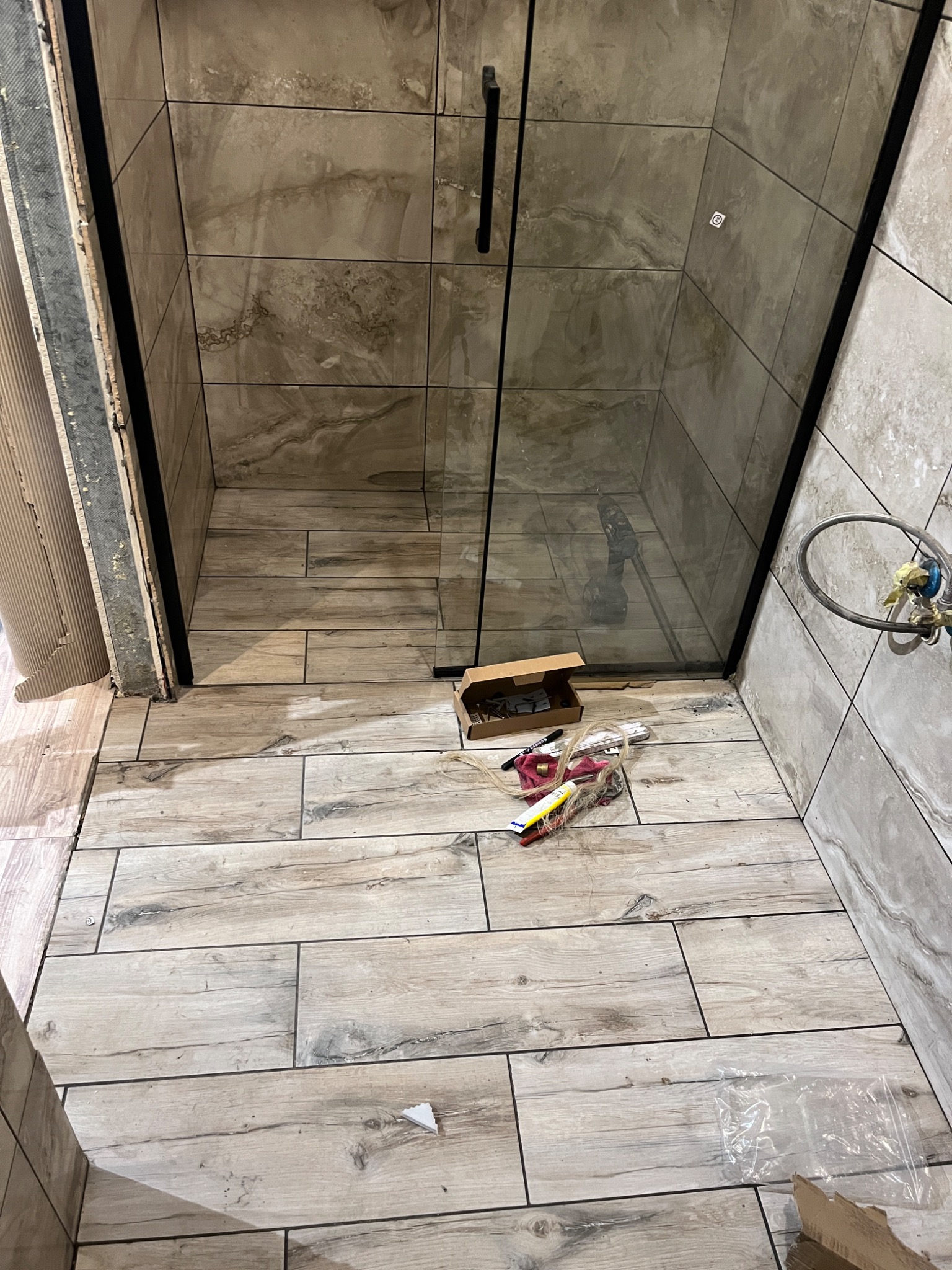 Płytki imitujące drewno na podłodze i ścianach kabiny prysznicowej w trakcie montażu. Widoczne narzędzia, kartonowe pudełko i niezakończone wykończenie przy wejściu do kabiny.