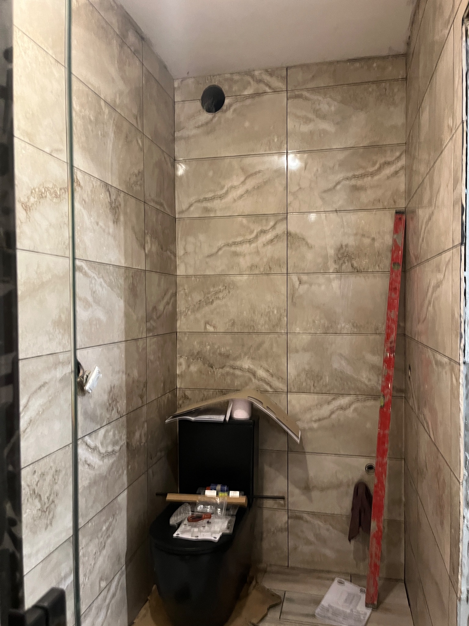 Łazienka w trakcie remontu: kremowe płytki imitujące marmur na ścianach i podłodze, czarna toaleta z deską i narzędziami na wierzchu, czerwona poziomica oparta o ścianę, widoczny otwór wentylacyjny...