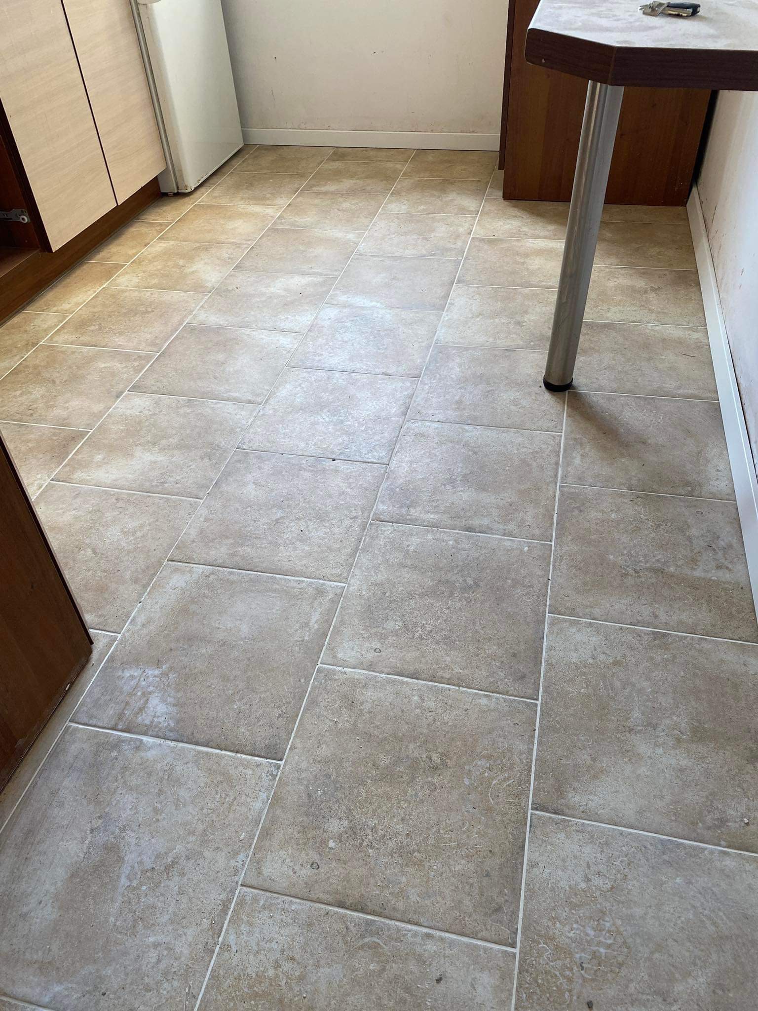 Beżowe, matowe płytki podłogowe ułożone w kuchni, z widocznymi fugami, fragmentem szafki kuchennej i nogą stołu w tle.