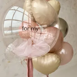 Kompozycja balonowa z transparentnym balonem wypełnionym tiulem i konfetti, z napisem 'for my beautiful', otoczona balonami w odcieniach różu i złota, na tle okna z widokiem na budynek.