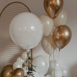 Fundacji Rozwoju Przedsiębiorczości Twój StartUp - Kompozycja z białych i złotych balonów, z dodatkiem zielonych gałązek, ustawiona na tle złotej, metalowej konstrukcji w kształcie łuku.