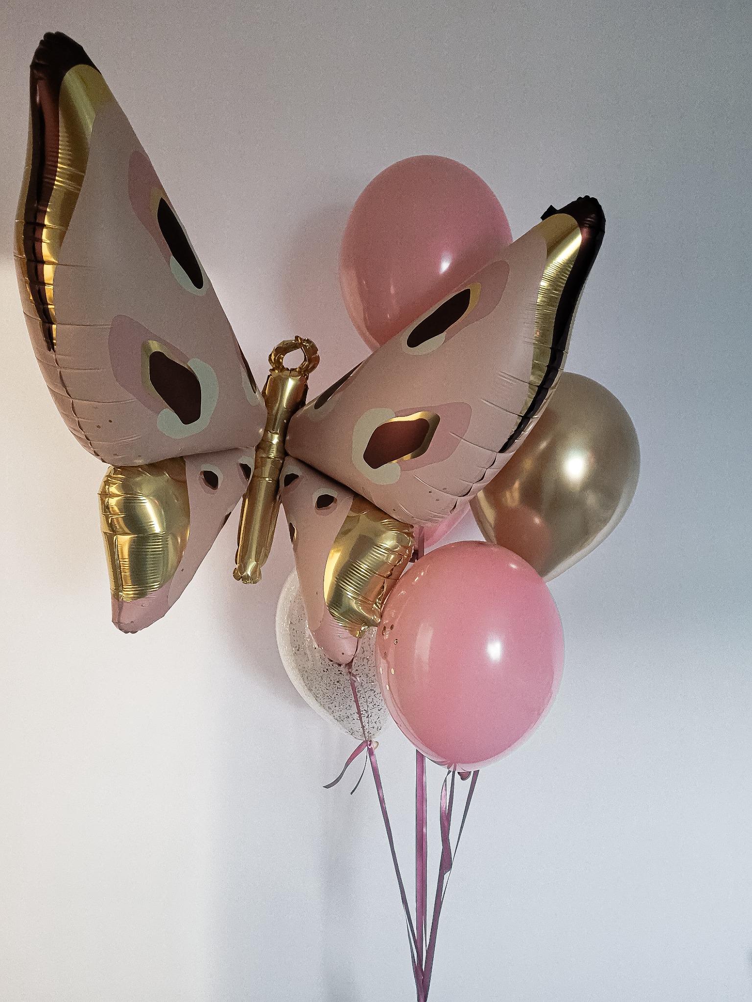 Bukiet balonów z dużym balonem w kształcie motyla w pastelowych kolorach z złotymi akcentami, uzupełniony o różowe i złote balony okrągłe, całość na tle białej ściany.