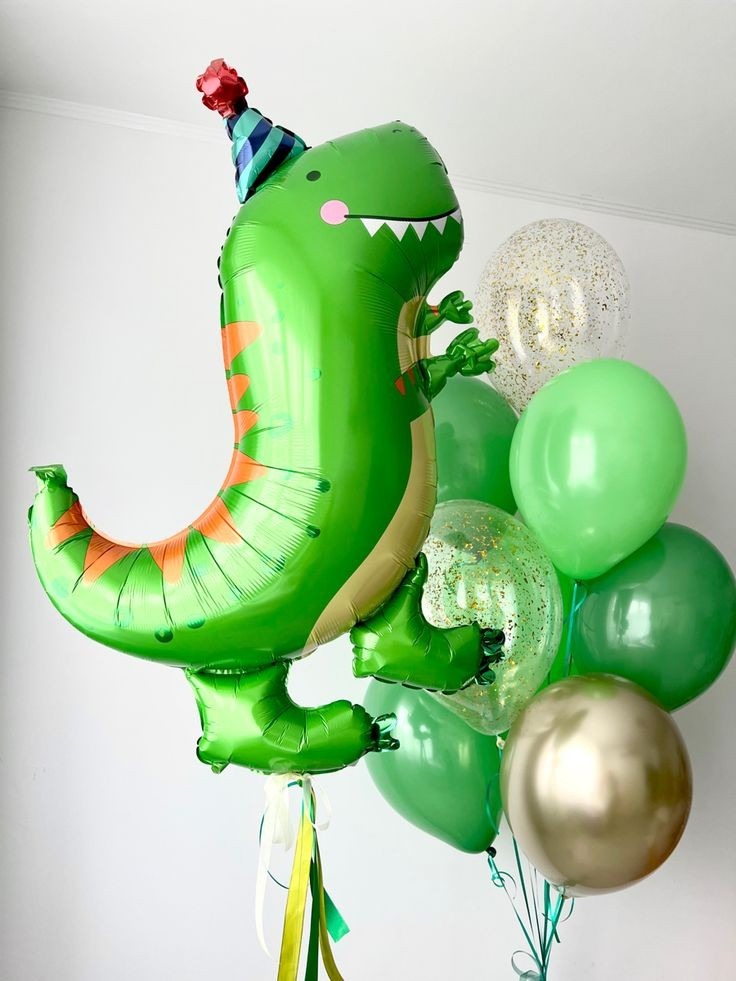 Kompozycja balonowa z dużym balonem foliowym w kształcie zielonego dinozaura w czapeczce urodzinowej, otoczonym przez balony lateksowe w odcieniach zieleni i przezroczyste balony z konfetti.