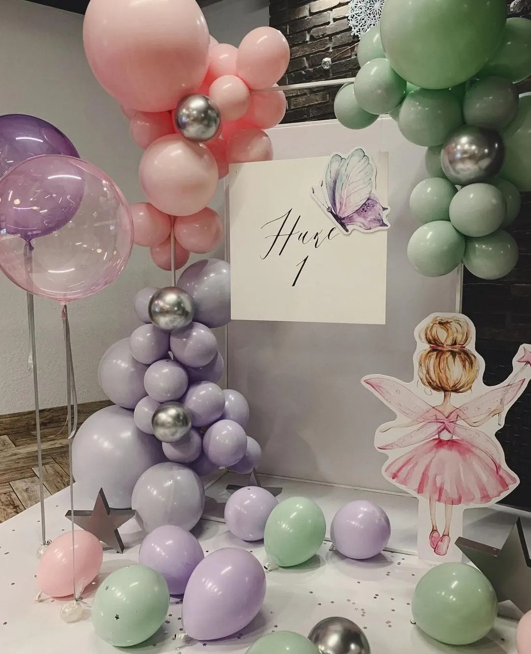 Dekoracja balonowa w pastelowych odcieniach różu, fioletu i zieleni z elementami srebrnymi, ustawiona na białym tle z kartonową figurką wróżki i napisem 'Hure 1' ozdobionym motylem, tworząca...