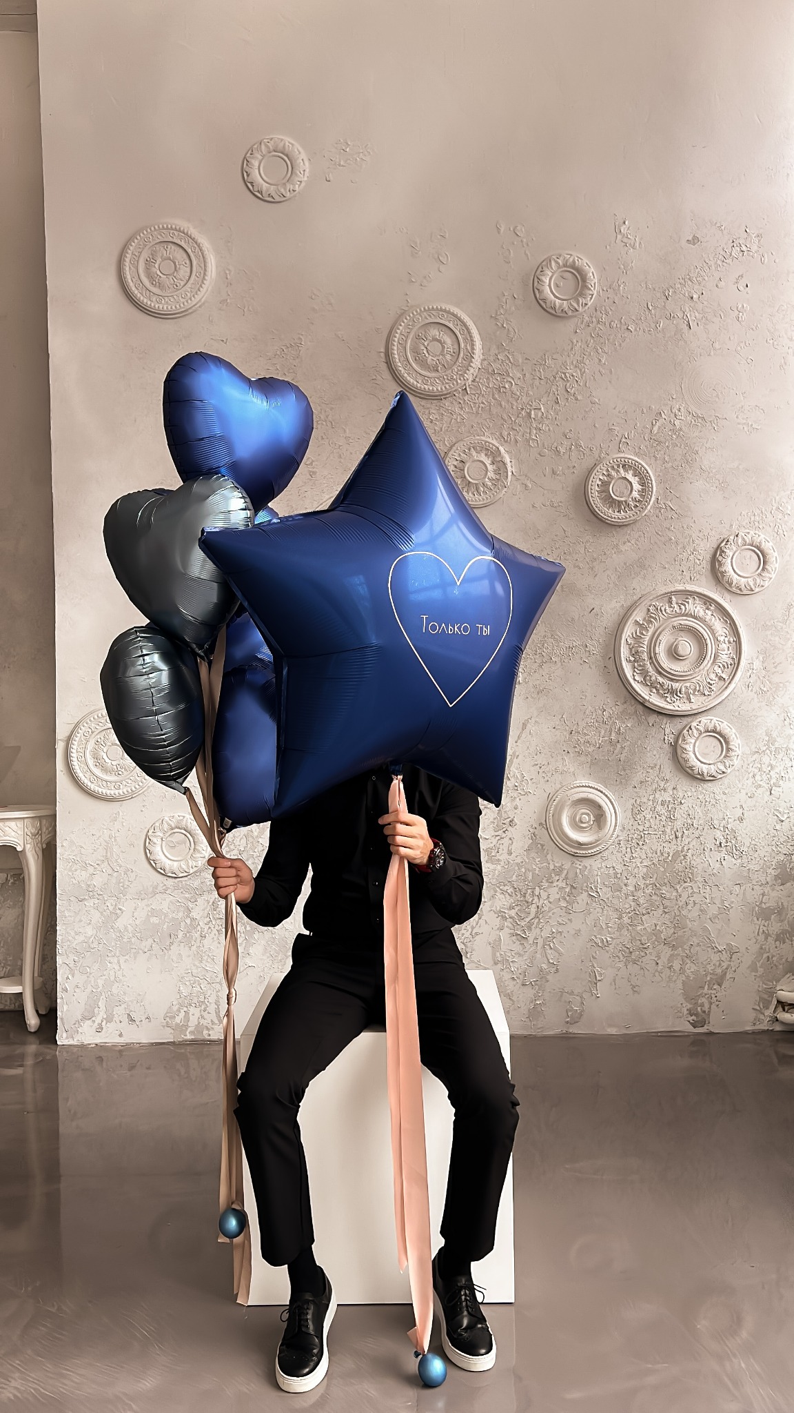 Osoba w czarnym stroju siedzi na białym sześcianie, zasłaniając twarz bukietem niebieskich balonów w kształcie serc i gwiazdy z napisem 'Только ты' w sercu, na tle ściany z dekoracyjnymi rozetami.