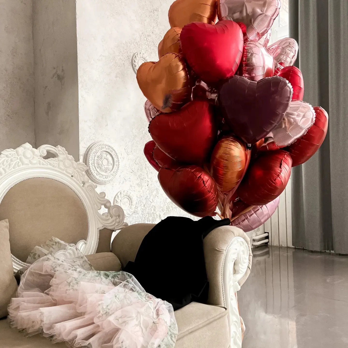 Kompozycja z balonów w kształcie serc w odcieniach czerwieni, różu i złota, ustawiona obok ozdobnego fotela z beżową tapicerką i leżącą na nim tiulową spódniczką w kwiaty oraz czarnym ubraniem,...