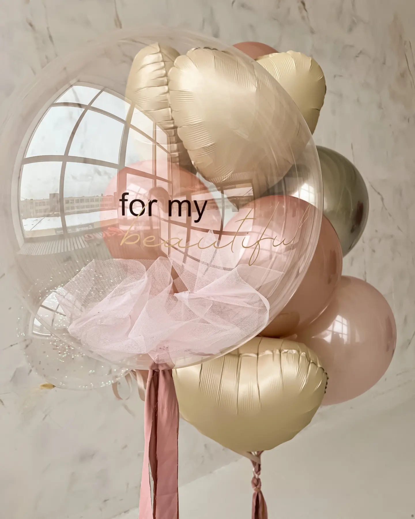Kompozycja balonowa z transparentnym balonem wypełnionym tiulem i konfetti, z napisem 'for my beautiful', otoczona balonami w odcieniach różu i złota, na tle okna z widokiem na budynek.
