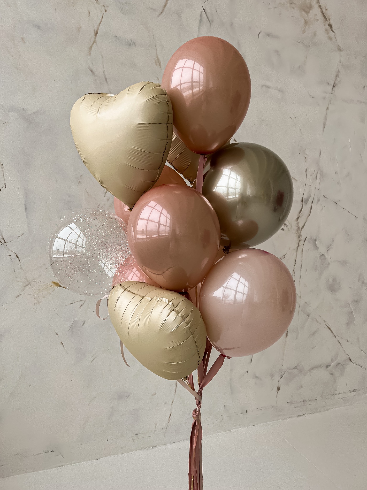 Bukiet balonów w odcieniach różu, beżu i złota, z balonem w kształcie serca i przezroczystym balonem z brokatem, zawiązany różową wstążką, na tle marmurowej ściany.