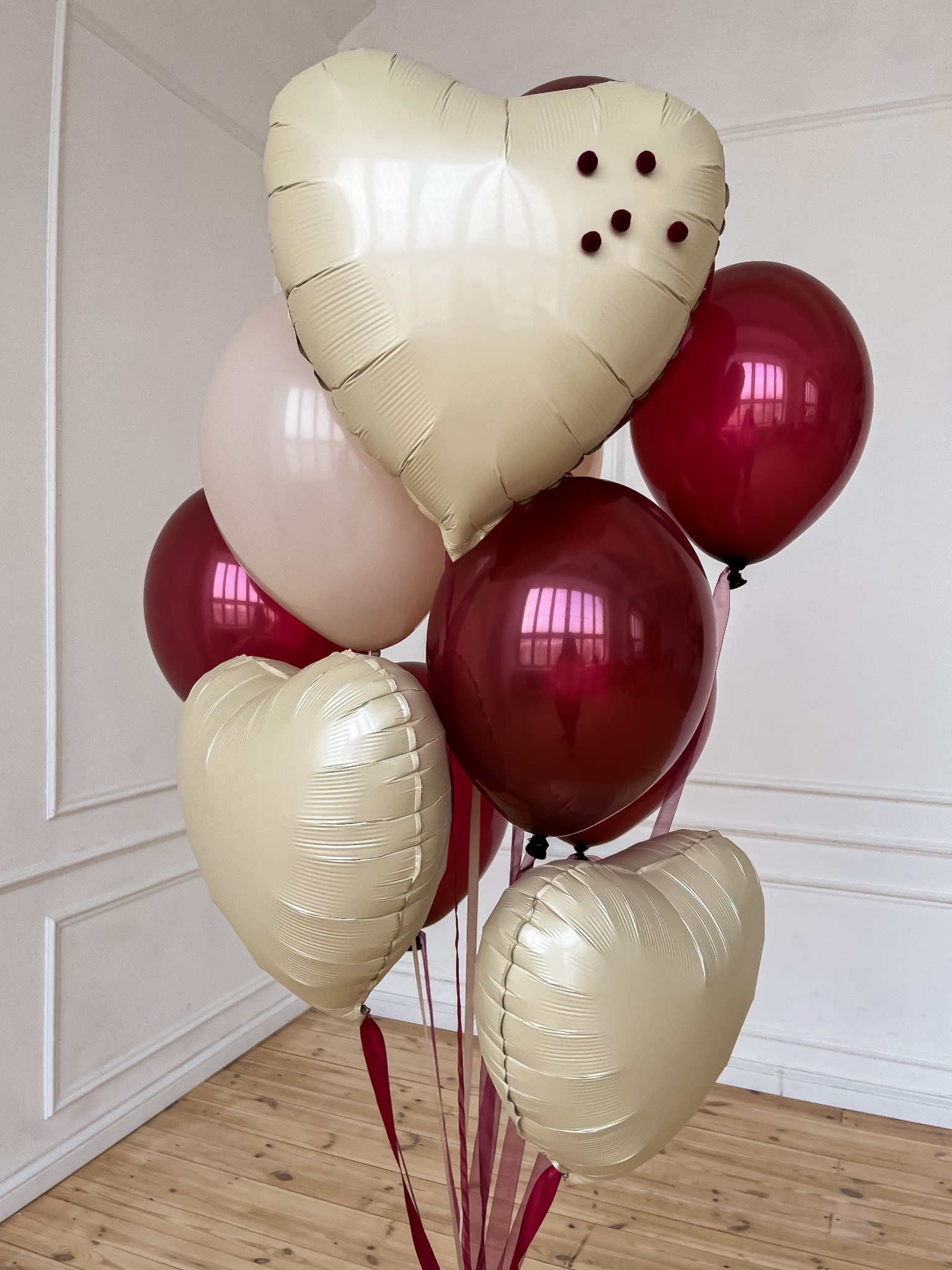 Bukiet balonów w kolorach czerwieni, beżu i pudrowego różu, z balonami w kształcie serc i okrągłymi, umieszczony na drewnianej podłodze, z dekoracyjnymi wstążkami.