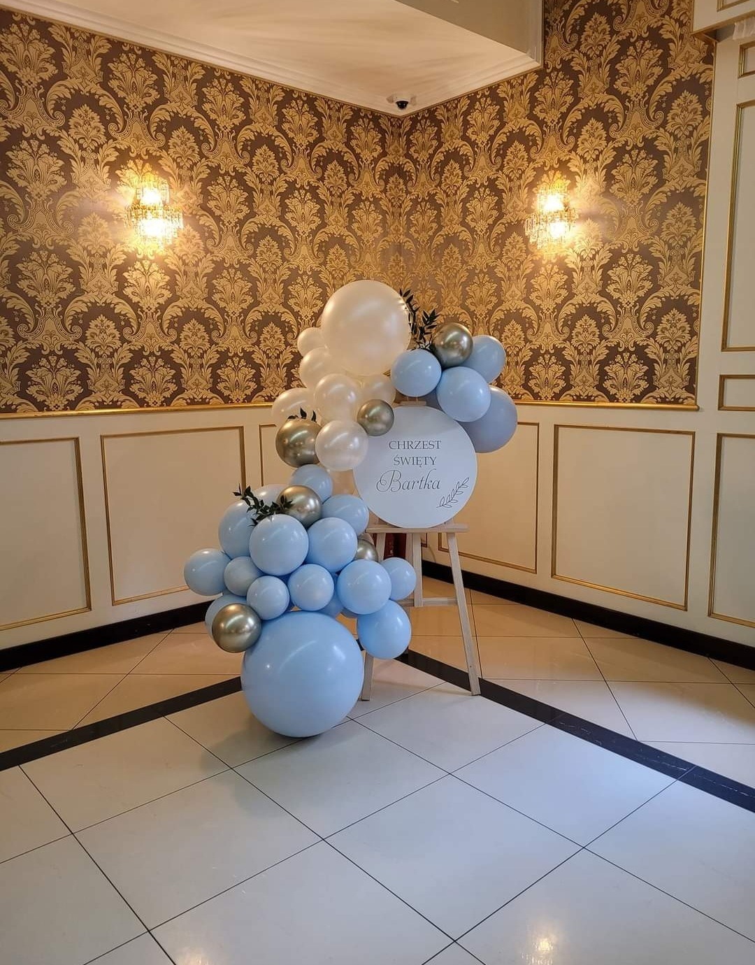 Dekoracja balonowa w odcieniach błękitu, bieli i złota z napisem 'Chrzest Święty Bartka' na drewnianym stojaku, ustawiona w eleganckim wnętrzu z wzorzystą tapetą.