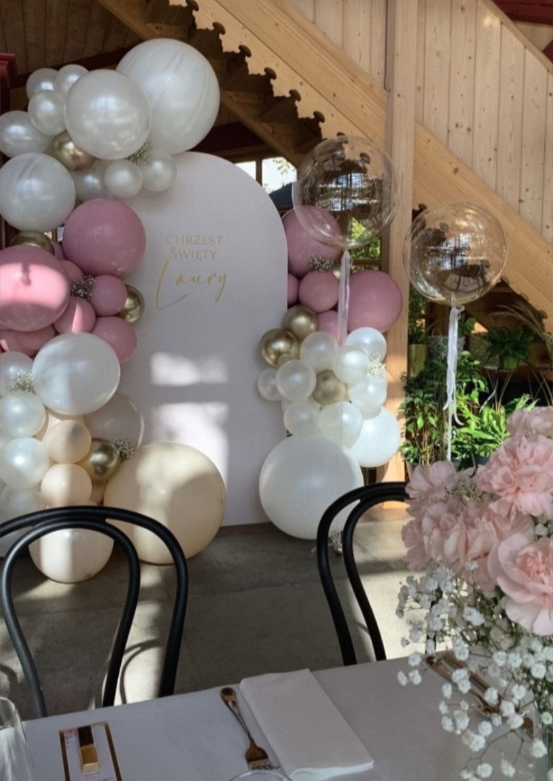 Dekoracja balonowa w kolorach białym, różowym i złotym z przezroczystymi balonami na Chrzest Święty Laury, ustawiona za stołem z nakryciem i bukietem kwiatów.