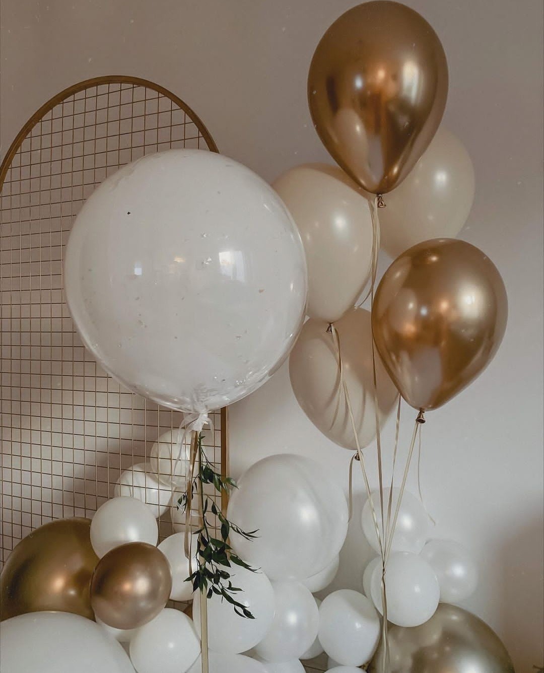 Kompozycja z białych i złotych balonów, z dodatkiem zielonych gałązek, ustawiona na tle złotej, metalowej konstrukcji w kształcie łuku.