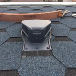 Szary odpowietrznik dachowy zamontowany na dachu pokrytym gontem bitumicznym w odcieniach niebieskiego i czarnego, obok leżą nożyce do blachy i kątownik dekarski, widoczna również miedziana obróbka...