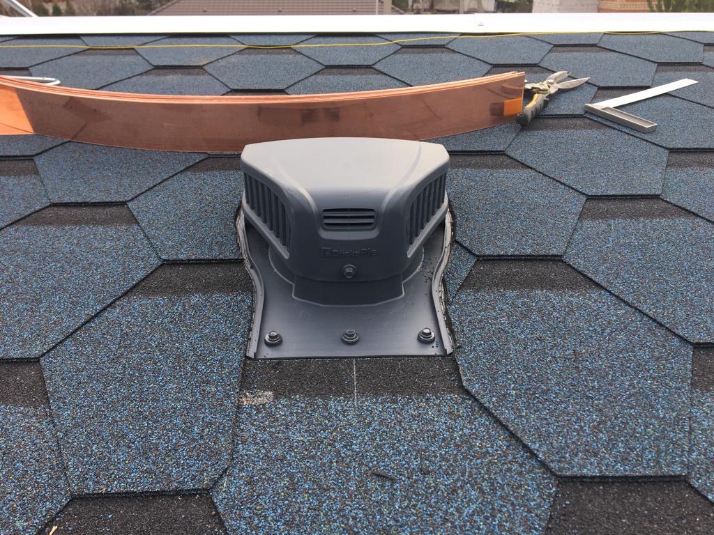 Szary odpowietrznik dachowy zamontowany na dachu pokrytym gontem bitumicznym w odcieniach niebieskiego i czarnego, obok leżą nożyce do blachy i kątownik dekarski, widoczna również miedziana obróbka...