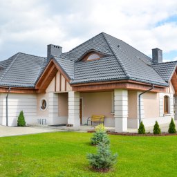 Budo-Montaż sp. z o.o. sp. komandytowa` - Nowoczesny dom jednorodzinny z beżową elewacją, grafitową dachówką i drewnianymi elementami wykończeniowymi, otoczony zielonym trawnikiem z niskimi iglakami.