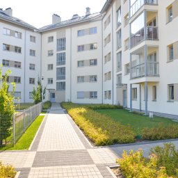 Budo-Montaż sp. z o.o. sp. komandytowa` - Nowoczesny budynek wielorodzinny z jasną elewacją, szklanymi balkonami, zadbanym trawnikiem i ścieżką z kostki brukowej w słoneczny dzień.