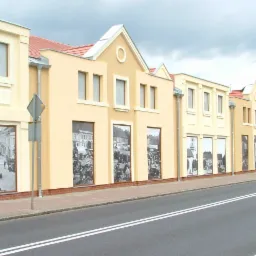 Fasada szeregu budynków pomalowanych na kremowo i żółto, z wkomponowanymi czarno-białymi fotografiami w stylu retro na parterze, widok z ulicy w pochmurny dzień.