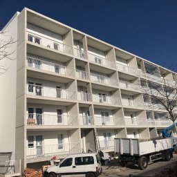 Budo-Montaż sp. z o.o. sp. komandytowa` - Wielopiętrowy budynek mieszkalny z balkonami, widoczna platforma robocza z pracownikiem, samochód dostawczy i materiały budowlane u podnóża budynku, błękitne niebo w tle.