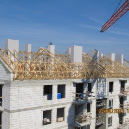 Budo-Montaż sp. z o.o. sp. komandytowa` - Budowa domu wielorodzinnego z białych bloczków, widoczne drewniane elementy konstrukcji dachu, kominy i fragment żurawia budowlanego na tle błękitnego nieba.