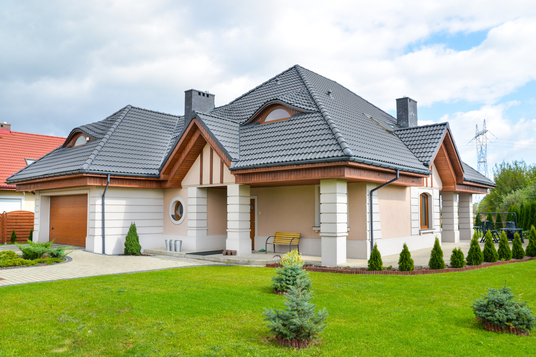 Nowoczesny dom jednorodzinny z beżową elewacją, grafitową dachówką i drewnianymi elementami wykończeniowymi, otoczony zielonym trawnikiem z niskimi iglakami.