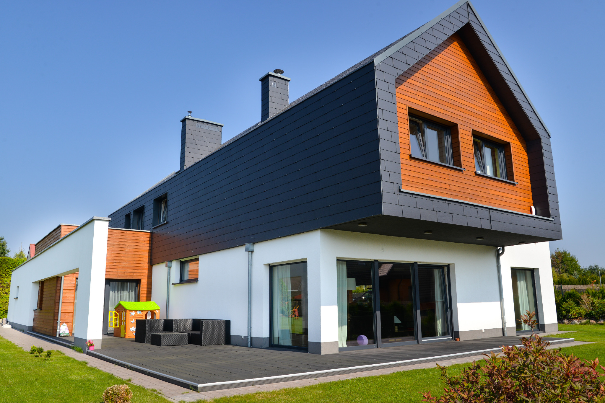 Nowoczesny dom z elewacją z drewnianych paneli i grafitowej dachówki, taras z meblami ogrodowymi i placem zabaw.